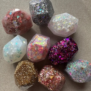 May include: A collection of ten small, faceted, resin gemstone-shaped ornaments. Each ornament is filled with glitter and iridescent flakes in various colors, including pink, gold, silver, and purple.