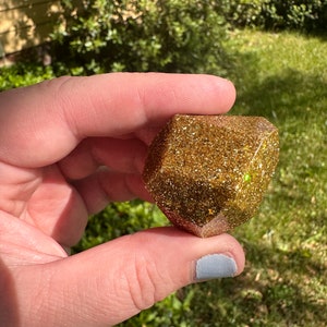 K&ouml;nnte beinhalten: Ein goldener Glitzer-Harzw&uuml;rfel mit einem facettenreichen Design. Der W&uuml;rfel wird in einer Hand vor einem Hintergrund aus gr&uuml;nem Laub und Sonnenlicht gehalten. Der W&uuml;rfel ist ein einzigartiger, handgefertigter Artikel.