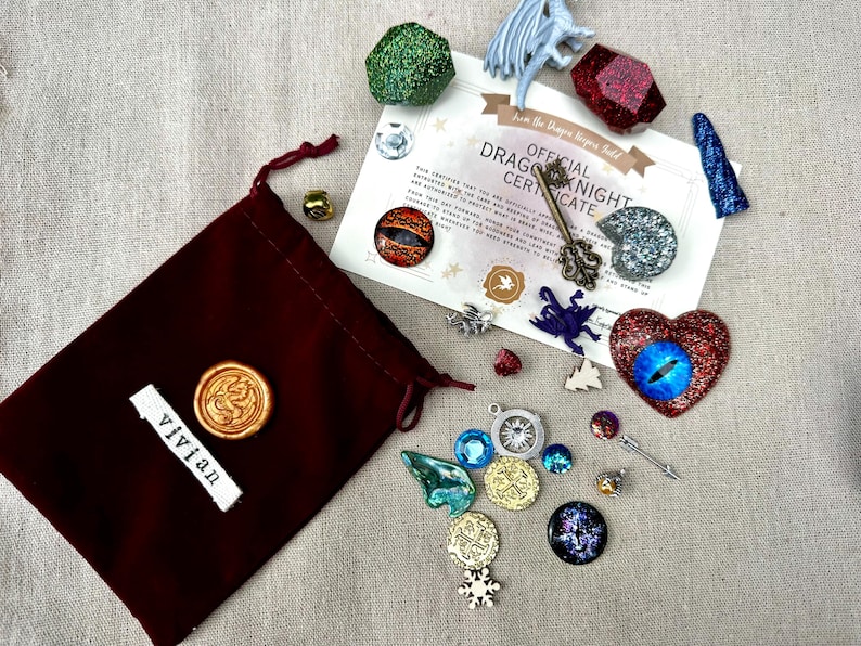 K&ouml;nnte beinhalten: Ein burgunderfarbener Samtbeutel mit einem goldenen Wachssiegel und einem Etikett mit der Aufschrift "vivian" neben verschiedenen Fantasy-Charms, Edelsteinen und einem Zertifikat. Das Zertifikat lautet "Official Dragon Knight Certificate".