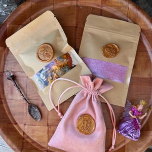 May include: An assortment of items on a wooden tray. Two kraft paper pouches sealed with gold wax stamps, a pink velvet drawstring bag with a wax seal, a decorative spoon, and a small fairy figurine.