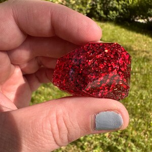 K&ouml;nnte beinhalten: Ein facettiertes, rechteckiges Objekt mit tiefrotem Glitzer, eingebettet in klarem Harz. Das Objekt wird in einer Hand vor einem verschwommenen gr&uuml;nen Hintergrund gehalten.