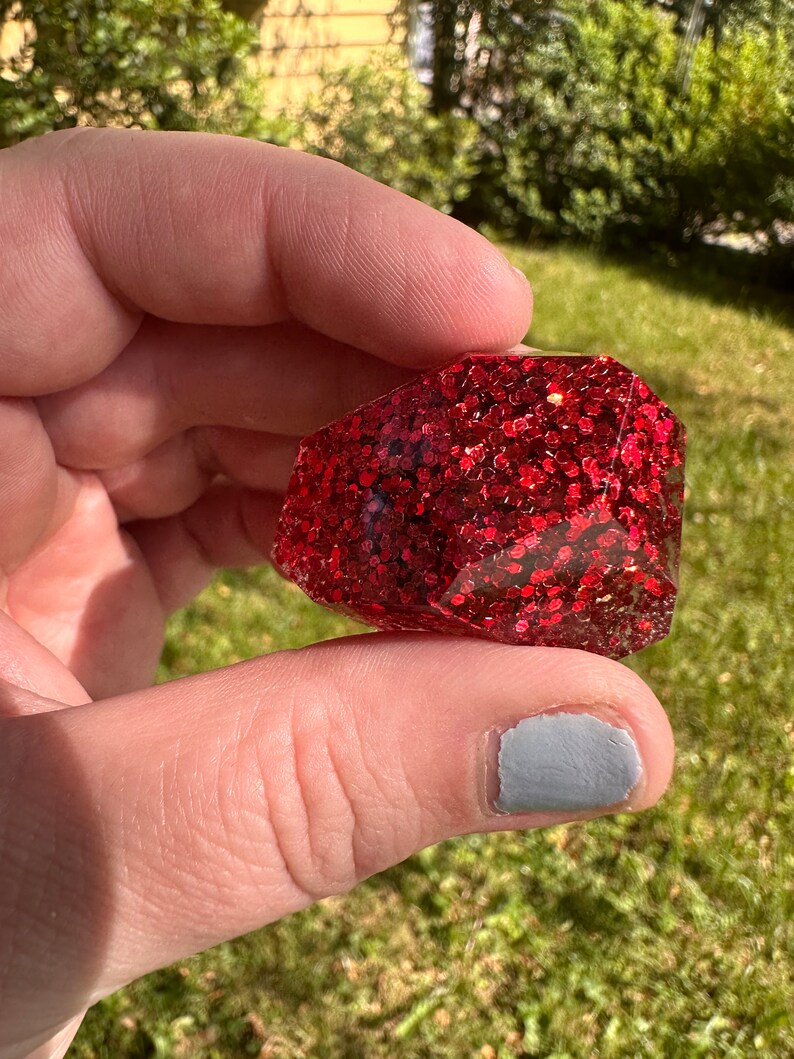 K&ouml;nnte beinhalten: Ein facettiertes, rechteckiges Objekt mit tiefrotem Glitzer, eingebettet in klarem Harz. Das Objekt wird in einer Hand vor einem verschwommenen gr&uuml;nen Hintergrund gehalten.