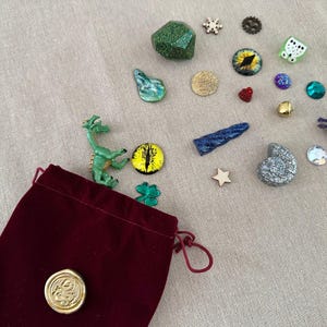 K&ouml;nnte beinhalten: Ein burgunderfarbener Samtbeutel mit Kordelzug und goldenem Siegel, umgeben von verschiedenen Fantasy-Miniaturen. Dazu geh&ouml;ren eine gr&uuml;ne Drachenfigur, ein glitzernder gr&uuml;ner W&uuml;rfel und verschiedene edelsteinartige Verzierungen in verschiedenen Farben und Formen.