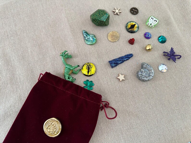 K&ouml;nnte beinhalten: Ein burgunderfarbener Samtbeutel mit Kordelzug und goldenem Siegel, umgeben von verschiedenen Fantasy-Miniaturen. Dazu geh&ouml;ren eine gr&uuml;ne Drachenfigur, ein glitzernder gr&uuml;ner W&uuml;rfel und verschiedene edelsteinartige Verzierungen in verschiedenen Farben und Formen.