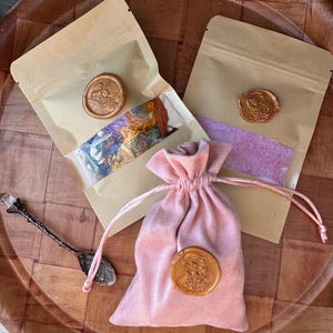 May include: An overhead shot of a wooden tray holding two sealed kraft paper pouches, a pink velvet drawstring bag, and a decorative spoon. The pouches are sealed with wax stamps. The bag also has a wax stamp. The pouches contain dried flowers and a purple powder.