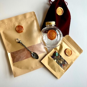 May include: A set including a clear glass sphere with a gold lid, a dark red velvet drawstring pouch, two tan paper pouches, and a metal spoon with a crystal handle. Each pouch and the glass sphere feature a circular gold wax seal.