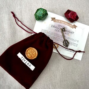 K&ouml;nnte beinhalten: Ein burgunderfarbener Samtbeutel mit Kordelzug, einem goldenen Wachssiegel und einem wei&szlig;en Etikett mit der Aufschrift "vivian". Ein Zertifikat mit dem Text "Official Dragon Knight Certificate" und ein dekorativer Schl&uuml;ssel sowie zwei facettierte Edelsteine.