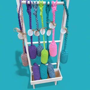 May include: A display of colorful crocheted dog toys hanging from a white wooden rack. The toys are made of different colored yarn and have a variety of shapes. The toys are labeled with the text "Woof! Dog Toy".