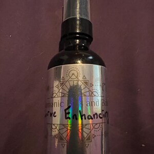May include: A black spray bottle with a clear cap. The silver label features geometric designs and the words "aromatic bliss and curve enhancing". The bottle is on a dark purple background.