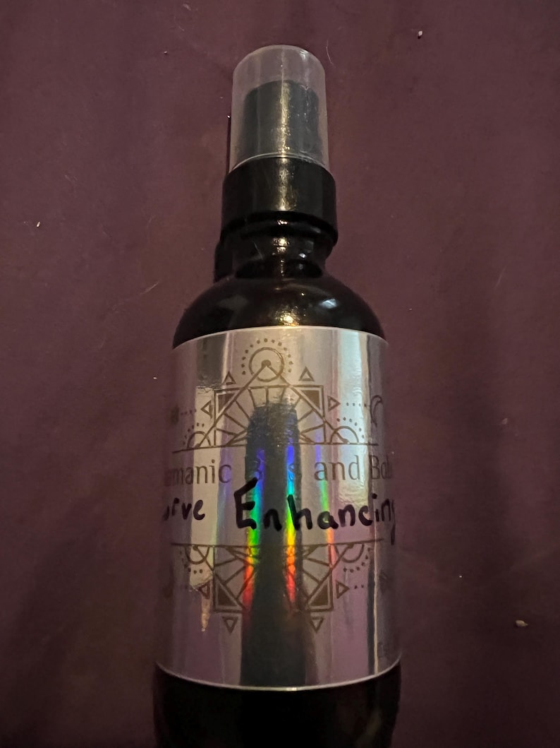 May include: A black spray bottle with a clear cap. The silver label features geometric designs and the words "aromatic bliss and curve enhancing". The bottle is on a dark purple background.