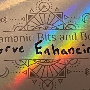 May include: A silver holographic surface with the text "Shamanic Bits and Bobs Curve Enhancing" in black. The design includes geometric shapes, crescent moons, and leaf-like elements. A rainbow sheen adds a colorful effect.