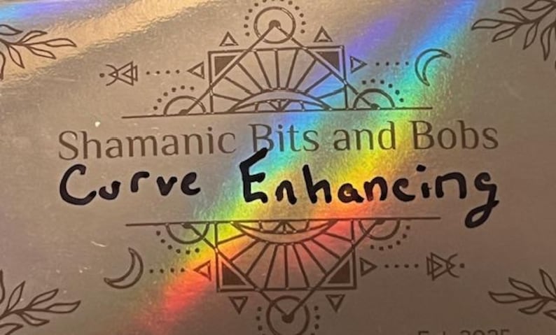 May include: A silver holographic surface with the text "Shamanic Bits and Bobs Curve Enhancing" in black. The design includes geometric shapes, crescent moons, and leaf-like elements. A rainbow sheen adds a colorful effect.