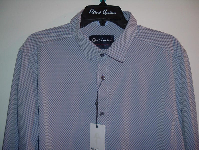 Puede incluir: Camisa azul claro con un peque&ntilde;o estampado de cuadros, colgada de una percha negra. El nombre de la marca "Robert Graham" est&aacute; impreso en la percha y una etiqueta. La camisa tiene cuello abotonado.