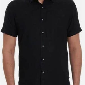 Puede incluir: Una camisa negra de manga corta con botones y cuello cl&aacute;sico. La camisa se abrocha con botones blancos. La tela parece ligera. La camisa la lleva una persona.
