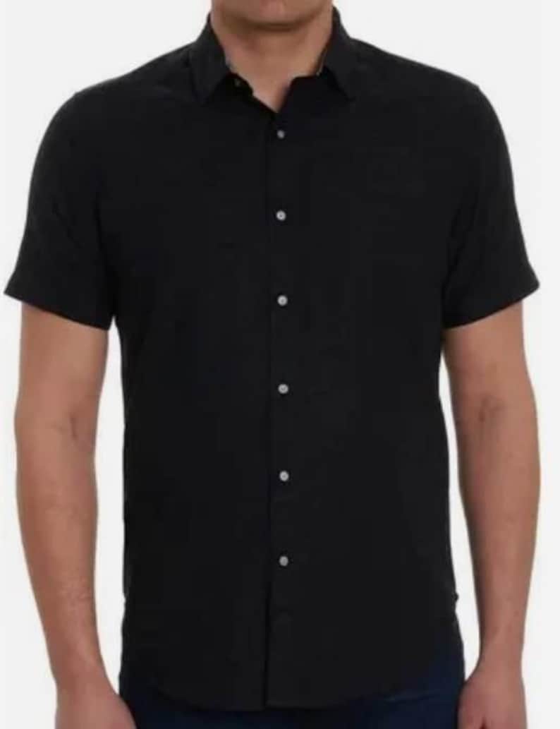 Puede incluir: Una camisa negra de manga corta con botones y cuello cl&aacute;sico. La camisa se abrocha con botones blancos. La tela parece ligera. La camisa la lleva una persona.