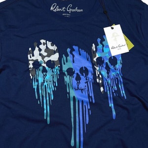 K&ouml;nnte beinhalten: Ein marineblaues T-Shirt mit einem grafischen Druck von drei tropfenden Sch&auml;deln in Blaut&ouml;nen und Gr&uuml;n. Die Sch&auml;del sind mit Tarnmustern gestaltet.