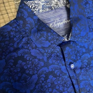 May include: Close-up of a blue button-down shirt with a dark floral pattern. The collar and cuffs have a contrasting white and blue design. The shirt has the brand name "Robert Graham" embroidered on the collar.