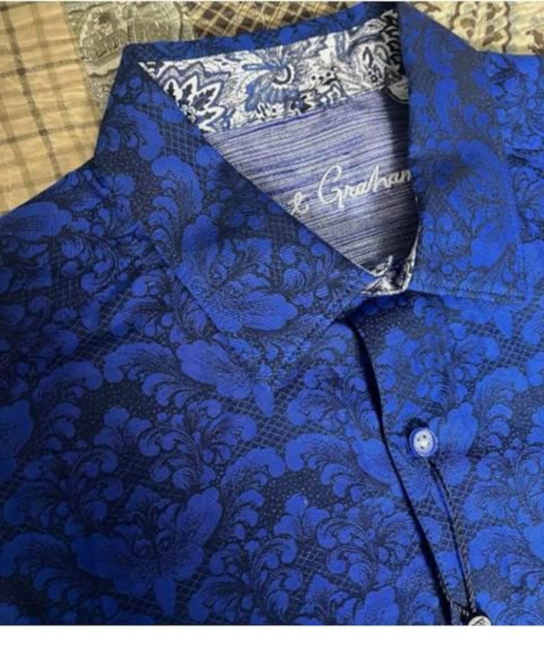 May include: Close-up of a blue button-down shirt with a dark floral pattern. The collar and cuffs have a contrasting white and blue design. The shirt has the brand name "Robert Graham" embroidered on the collar.