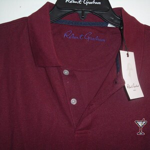May include: A burgundy polo shirt with a collar and three-button placket. The shirt features the embroidered text "Robert Graham" in blue on the chest and a small cocktail glass logo. A white tag with the brand name hangs from the collar.