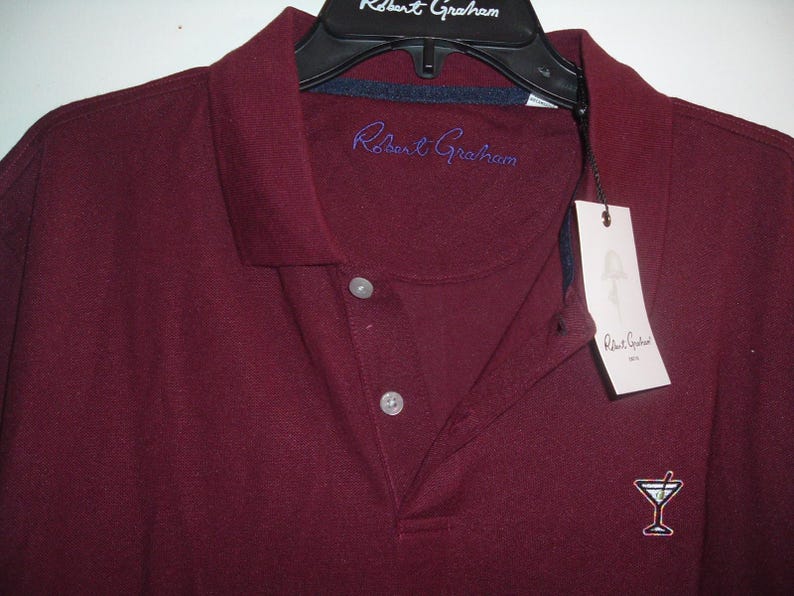 May include: A burgundy polo shirt with a collar and three-button placket. The shirt features the embroidered text "Robert Graham" in blue on the chest and a small cocktail glass logo. A white tag with the brand name hangs from the collar.