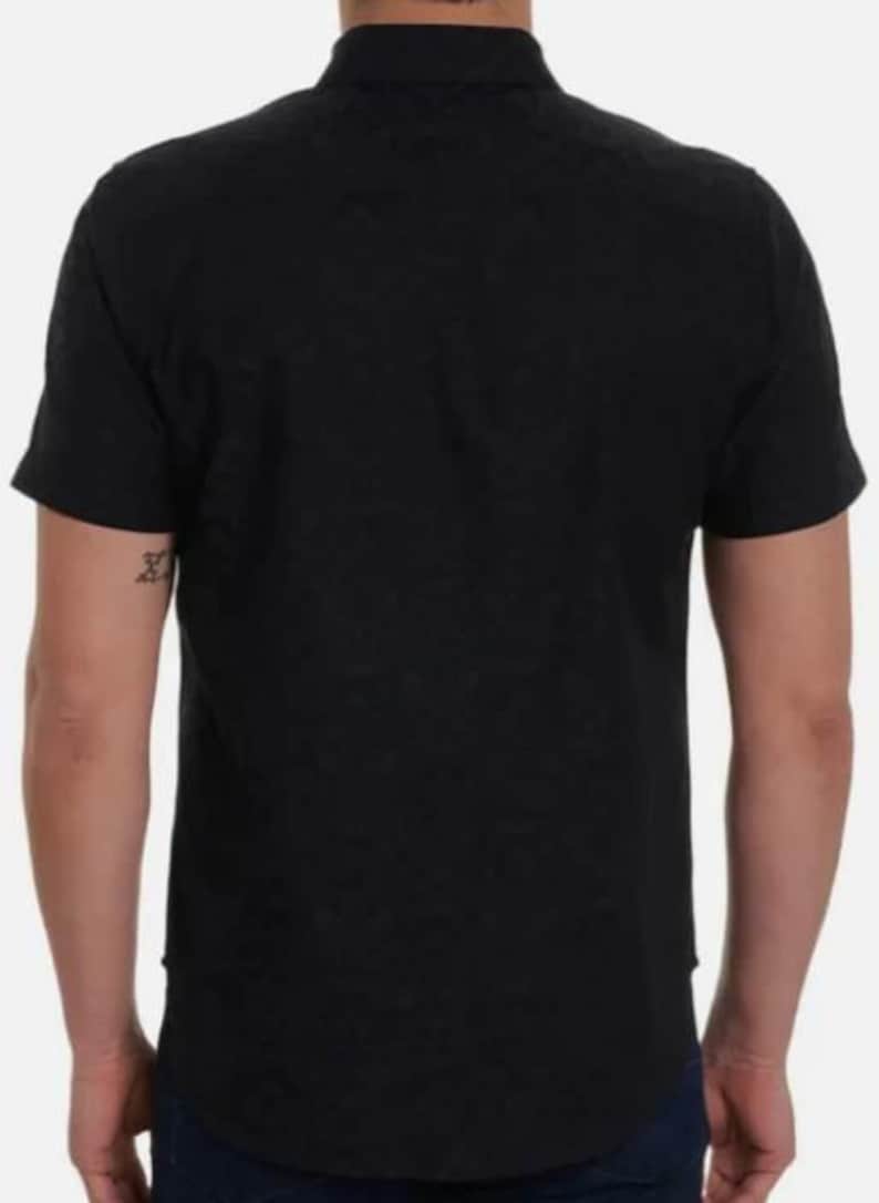 Puede incluir: Una camisa negra de manga corta con botones. La camisa tiene un cuello cl&aacute;sico y est&aacute; hecha de una tela oscura lisa. La vista trasera muestra el dise&ntilde;o y el ajuste de la camisa. El modelo lleva vaqueros azul oscuro.