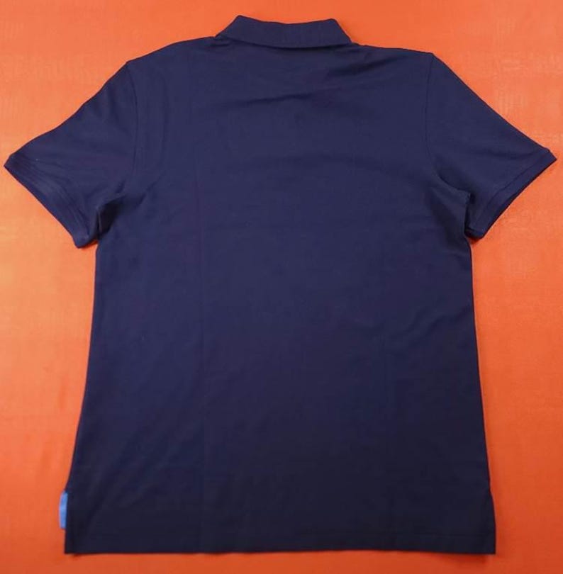 Puede incluir: Polo azul marino con mangas cortas y cuello. La camisa tiene un dise&ntilde;o cl&aacute;sico y est&aacute; hecha de una tela suave. Una peque&ntilde;a etiqueta azul es visible en la parte inferior izquierda del dobladillo.