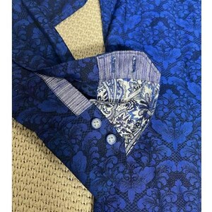 May include: Close-up of a blue button-down shirt with a dark blue floral pattern. The collar and cuffs feature a contrasting paisley design. The shirt has three blue buttons.