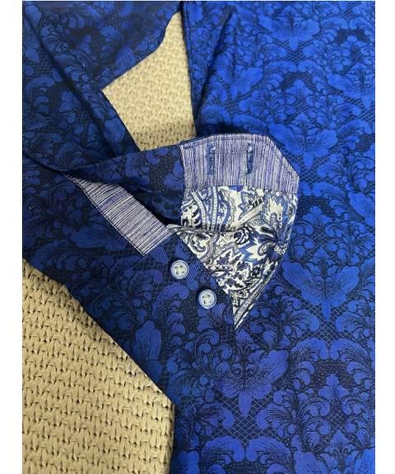 May include: Close-up of a blue button-down shirt with a dark blue floral pattern. The collar and cuffs feature a contrasting paisley design. The shirt has three blue buttons.