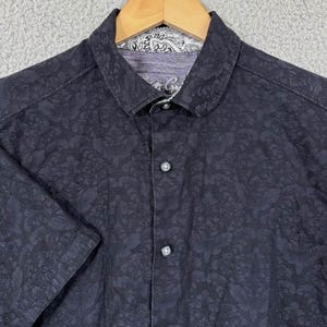 Puede incluir: Camisa azul marino oscuro de manga corta con estampado floral. La camisa tiene cuello y cierre de botones en la parte delantera. El forro del cuello tiene un dise&ntilde;o de cachemira. La camisa est&aacute; colgada en una percha de madera.