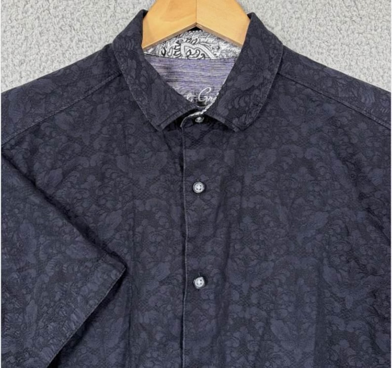 Puede incluir: Camisa azul marino oscuro de manga corta con estampado floral. La camisa tiene cuello y cierre de botones en la parte delantera. El forro del cuello tiene un dise&ntilde;o de cachemira. La camisa est&aacute; colgada en una percha de madera.