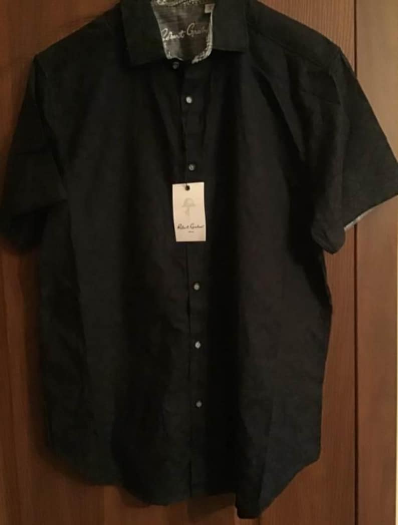 Puede incluir: Una camisa negra de manga corta con cuello y etiqueta. La camisa tiene una apariencia texturizada y una tela estampada en contraste en el interior del cuello y los pu&ntilde;os. La etiqueta dice "Robert Graham".