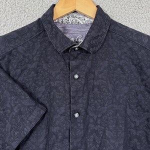 Puede incluir: Camisa abotonada de manga corta azul marino oscuro con un sutil estampado floral. La camisa presenta un cuello cl&aacute;sico y un estampado de cachemira en contraste en el cuello interior. Los botones son oscuros y la camisa est&aacute; en una percha de madera.