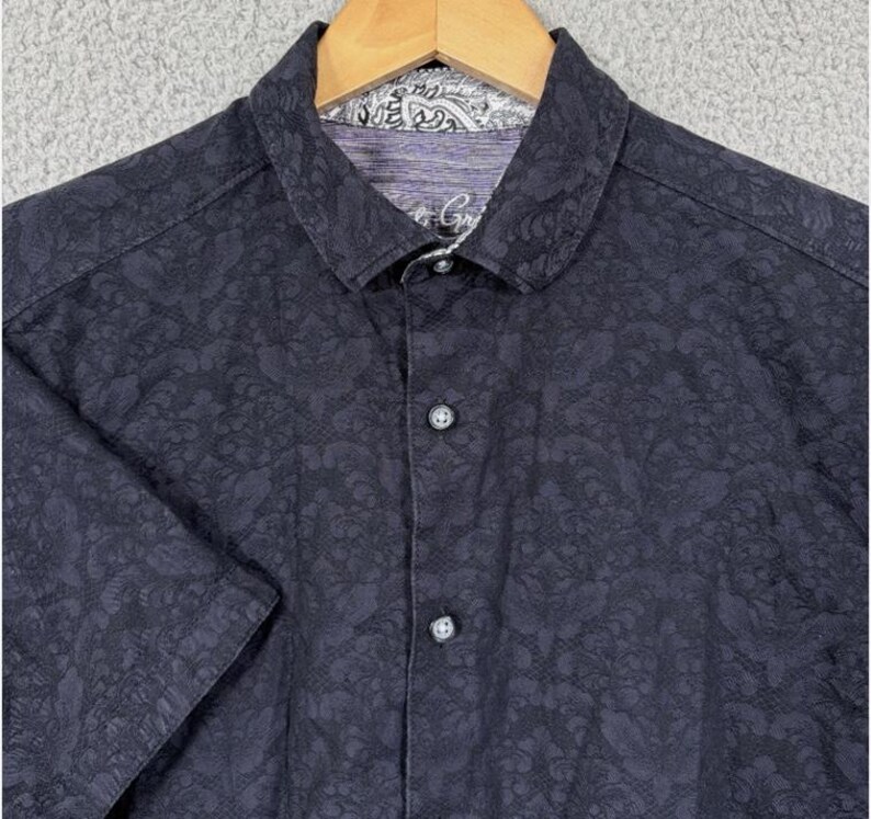 Puede incluir: Camisa abotonada de manga corta azul marino oscuro con un sutil estampado floral. La camisa presenta un cuello cl&aacute;sico y un estampado de cachemira en contraste en el cuello interior. Los botones son oscuros y la camisa est&aacute; en una percha de madera.