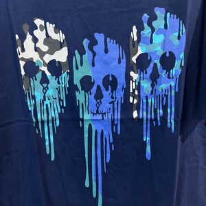 K&ouml;nnte beinhalten: Ein marineblaues T-Shirt mit drei tropfenden Sch&auml;delmotiven. Die Sch&auml;del sind in einem Tarnmuster mit blauen, gr&uuml;nen und wei&szlig;en Farben gehalten.