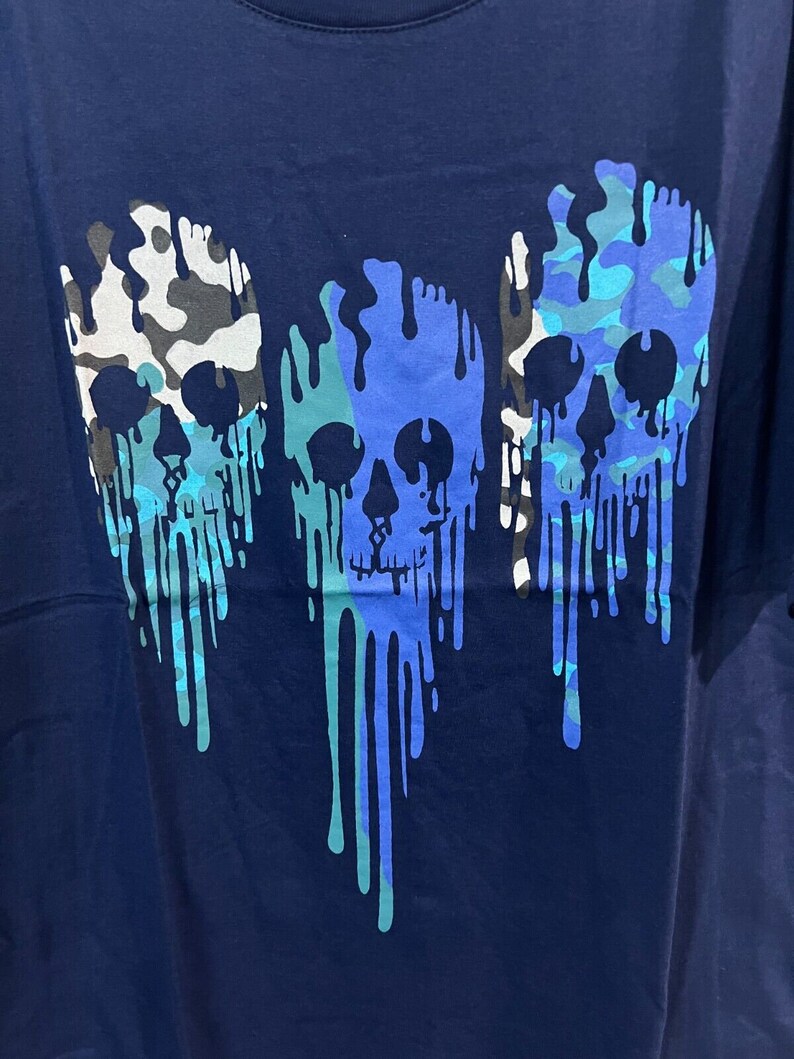 K&ouml;nnte beinhalten: Ein marineblaues T-Shirt mit drei tropfenden Sch&auml;delmotiven. Die Sch&auml;del sind in einem Tarnmuster mit blauen, gr&uuml;nen und wei&szlig;en Farben gehalten.