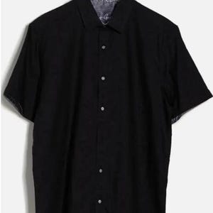 Puede incluir: Una camisa negra de manga corta con cuello, colgada de una percha de madera. La camisa tiene un patr&oacute;n sutil en el interior del cuello y los pu&ntilde;os. Est&aacute; abotonada y parece estar hecha de una tela ligera.