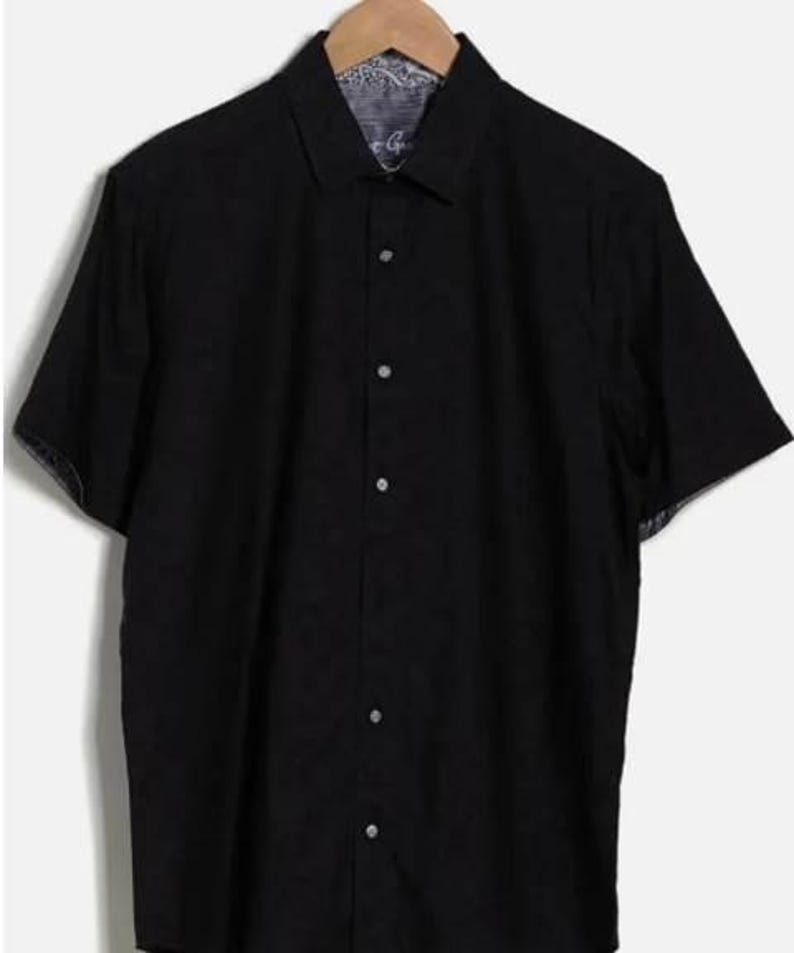 Puede incluir: Una camisa negra de manga corta con cuello, colgada de una percha de madera. La camisa tiene un patr&oacute;n sutil en el interior del cuello y los pu&ntilde;os. Est&aacute; abotonada y parece estar hecha de una tela ligera.