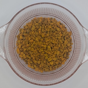 May include: A clear glass bowl filled with small, irregularly shaped pieces of dried, yellow-orange turmeric. The bowl has a slightly pinkish tint and a ridged design around the rim. The turmeric pieces are densely packed.