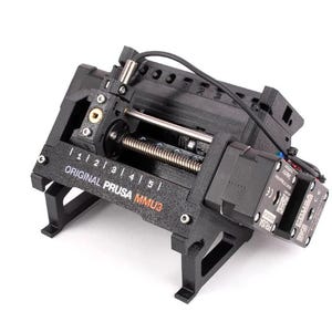 Könnte beinhalten: Ein schwarzer 3D-Drucker-Filament-Zuführer mit einem silbernen Zahnrad und einem schwarzen und orangefarbenen Etikett mit der Aufschrift "ORIGINAL PRUSA MMU3".