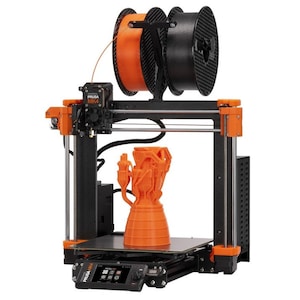 May include: A black and orange 3D printer with a spool of orange filament and a spool of black filament. The printer is printing a small orange object that looks like a rocket engine.
