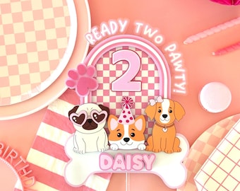 Puede incluir: Un adorno para pastel rosa y blanco con el texto "READY TWO PAWTY!" y el número "2". El adorno presenta tres perros de dibujos animados y el nombre "DAISY" en un hueso. Incluye una huella de pata rosa.