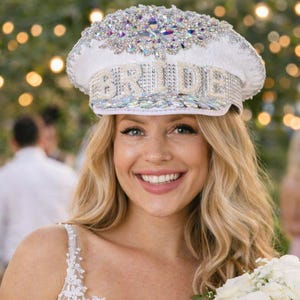 May include: A bride wearing a white cap with the word "BRIDE" in silver letters, adorned with rhinestones. She holds a bouquet of white and pink roses. The background features blurred figures and string lights, suggesting an outdoor wedding.