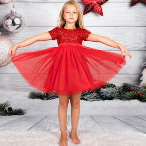 May include: A young girl wearing a red tulle dress with sequins. The dress has a fitted bodice and a full skirt. The girl is standing on a white fluffy rug in front of a wooden background.