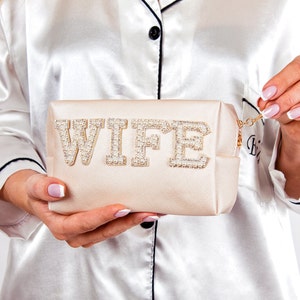 May include: A light beige satin makeup bag with a zipper closure. The bag has the word "WIFE" spelled out in large, white letters with pearl embellishments.