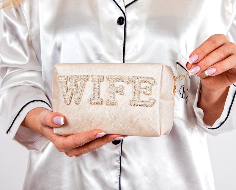 May include: A light beige satin makeup bag with a zipper closure. The bag has the word "WIFE" spelled out in large, white letters with pearl embellishments.