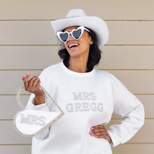 May include: A white sweatshirt with the text "MRS GREGG" in pearl-like embellishments. The person is wearing a white cowboy hat and heart-shaped sunglasses. They are holding a white heart-shaped purse with pearl-like embellishments and the text "MRS" on it.