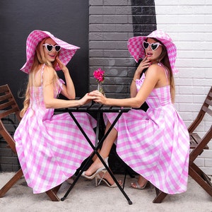 Puede incluir: Dos mujeres con vestidos a cuadros rosas y blancos a juego, sombreros de sol de gran tama&ntilde;o y gafas de sol en forma de coraz&oacute;n est&aacute;n sentadas en una peque&ntilde;a mesa negra. Se toman de la mano, con un peque&ntilde;o jarr&oacute;n de flores rosas entre ellas.