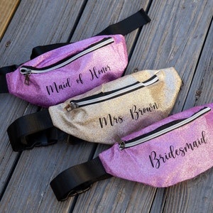 May include: Three sparkly fanny packs with black straps. The pink fanny pack says "Bridemaid", the gold fanny pack says "Mrs. Brown", and the purple fanny pack says "Maid of Honor".
