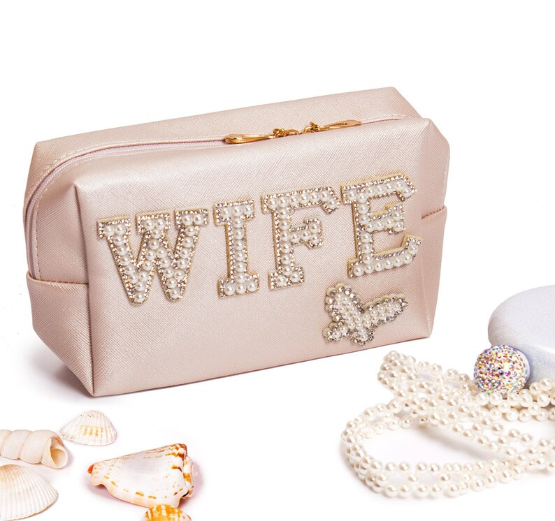 May include: A light pink zippered pouch with the word "WIFE" spelled out in pearl-like beads. The pouch has a gold zipper and a small pearl-like butterfly design.