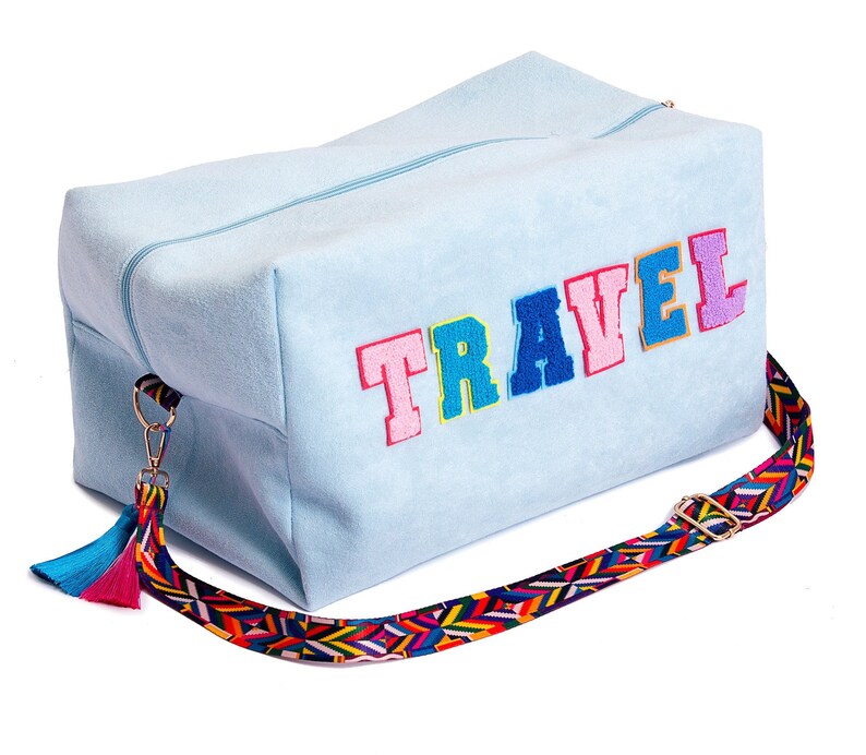 Puede incluir: Una bolsa de viaje azul claro con una aplicaci&oacute;n de la palabra "TRAVEL" de colores y una correa multicolor con un borla.